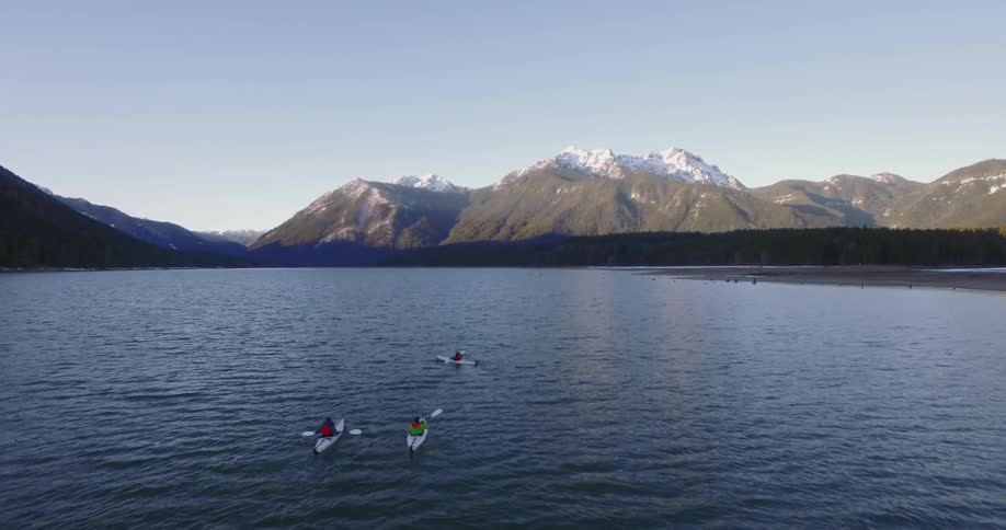 Three kayakers paddle on a body of water with snow-capped mountains in the background