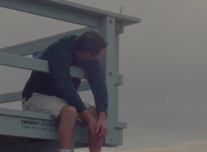 A man sits with his legs and arms dangling from the side of a lifeguard tower