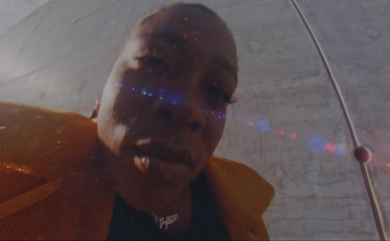 Black woman wearing a yellow overcoat dancing outdoors in front of a concrete wall