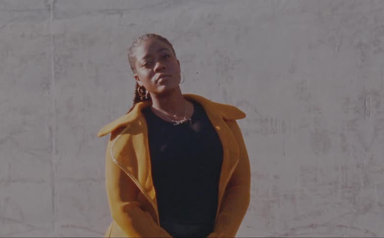 Woman with dreadlocks hoop earrings yellow overcoat and black shirt poses for the camera