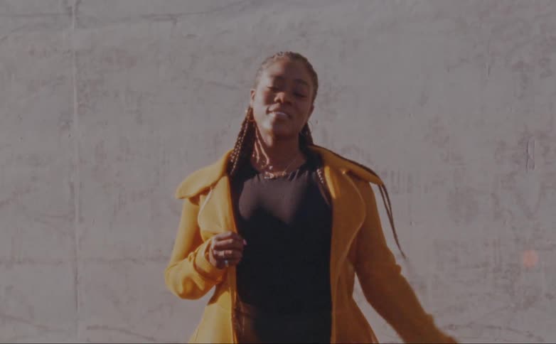Woman with dreadlocks hoop earrings yellow overcoat and black shirt poses for the camera