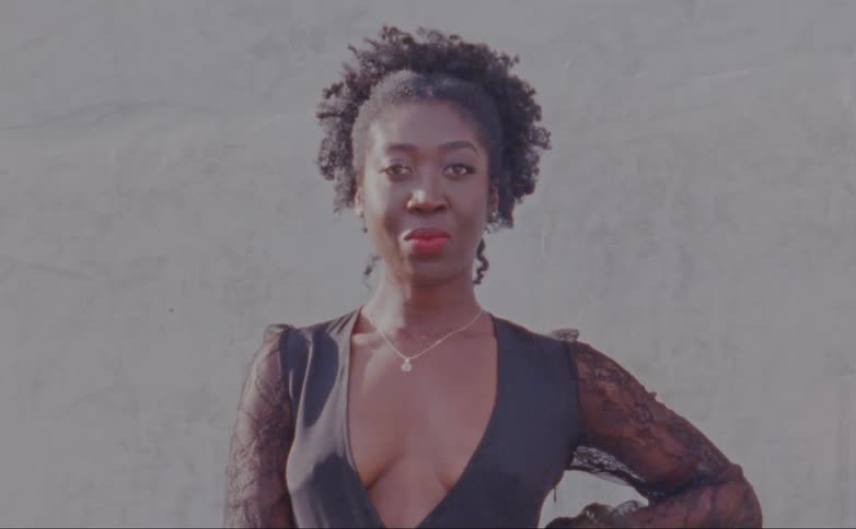 Female model with curly hair necklace and decollete dress poses for the camera standing in front of wall