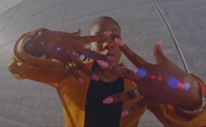 Black woman wearing a yellow overcoat showing her hands and dancing in front of a concrete wall outdoors
