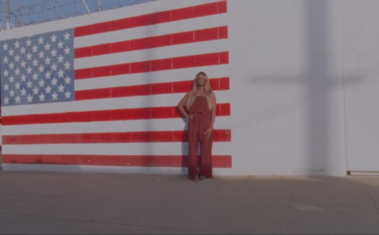 Woman with long blond hair stands in front of a large american flag
