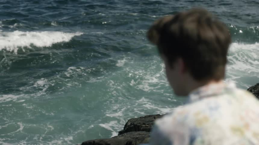 A young man sits on the shoreline cliffs as the waves crash
