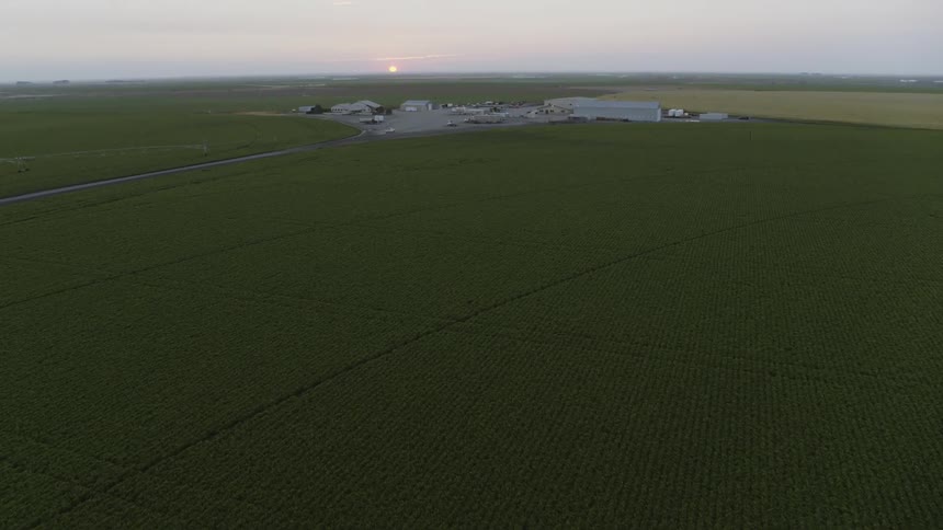 A truck drives into a farm yard between fields of crops as the morning sun sits just above the horizon