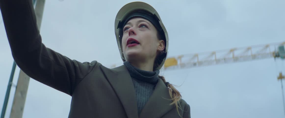 White woman wearing a white safety helmet gloves and a overcoat talking to someone and pointing at something at a construction site