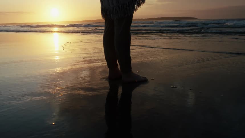 Woman walking towards the sea and stading there alone at an empty beach during sunset