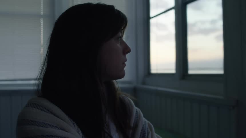 Woman sits in room wearing shawl pondering then looks toward window showing water and shore