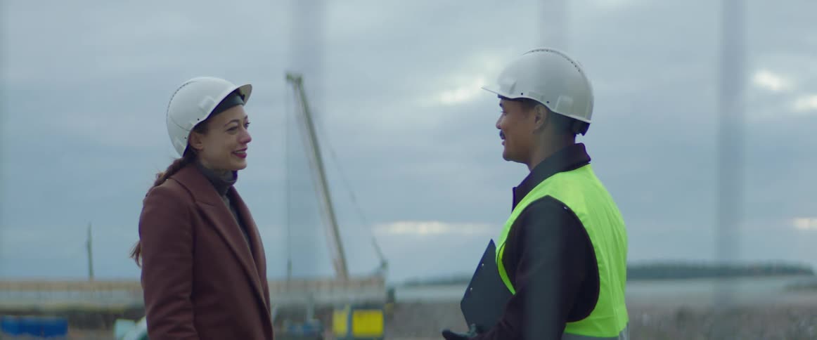 Woman wearing overcoat and safety helmet shaking the hand and talking to a construction worker wearing a green vest and safety helmet at a construction site