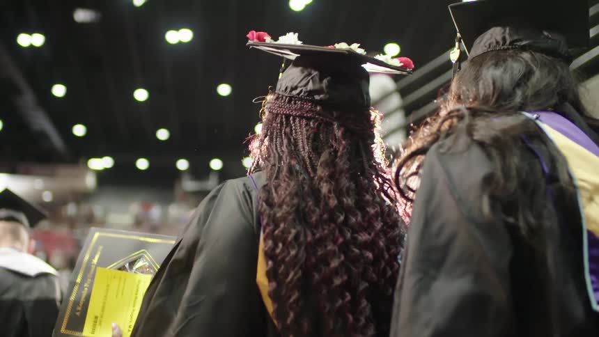Students are standing wearing caps and gowns at an indoor graduation ceremony in a stadium