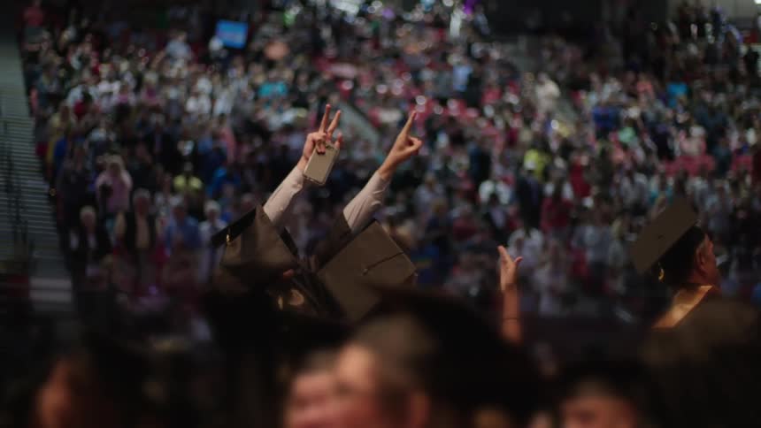Graduating students stand and cheer as their friends and family applause in the bleachers