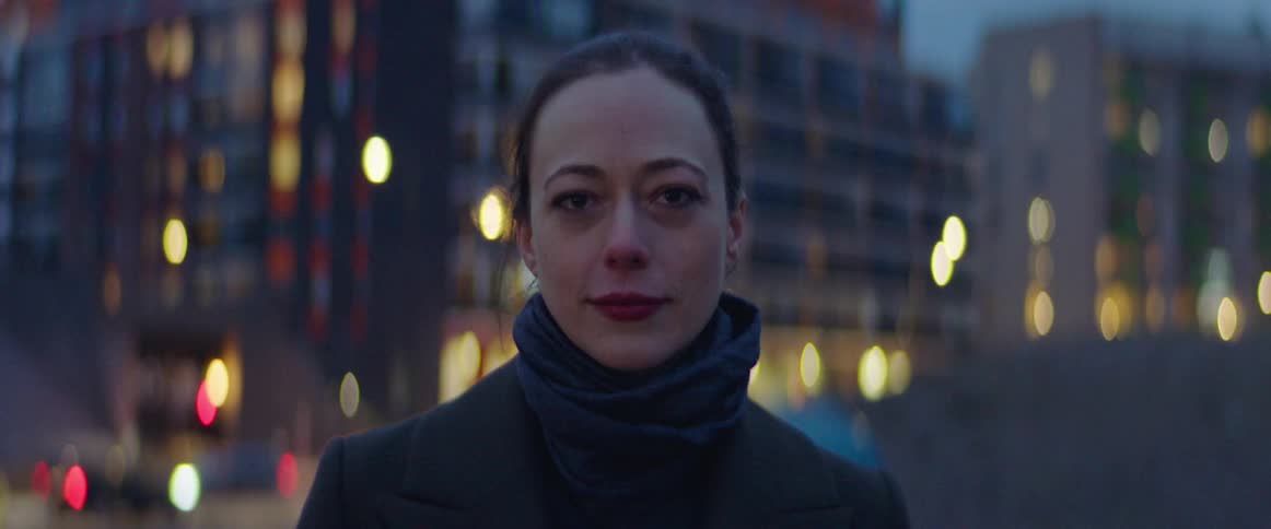 Woman with scarf and black coat faces the camera and cries tears of joy in the street with tall buildings in the background at dusk
