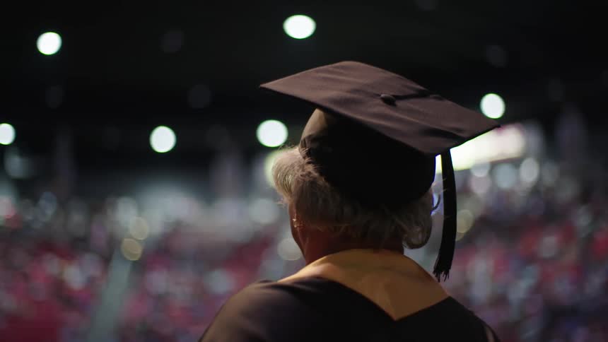 An elderly black woman wearing a cap and gown walks on stage in front of a large crowd