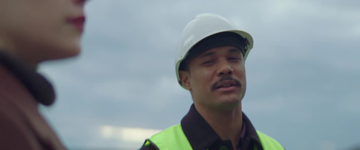 Man in a safety vest and hard hat talks to a woman while standing outdoors under a cloudy sky