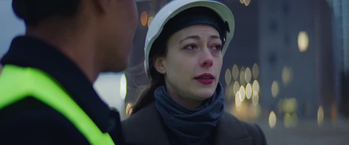 White woman wearing a scarf and safety helmet talking to a male construction worker wearing green vest and safety helmet