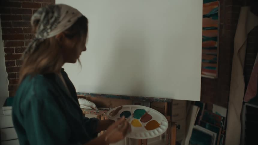 A young female artist mixes paint in a palette in front of the blank canvas