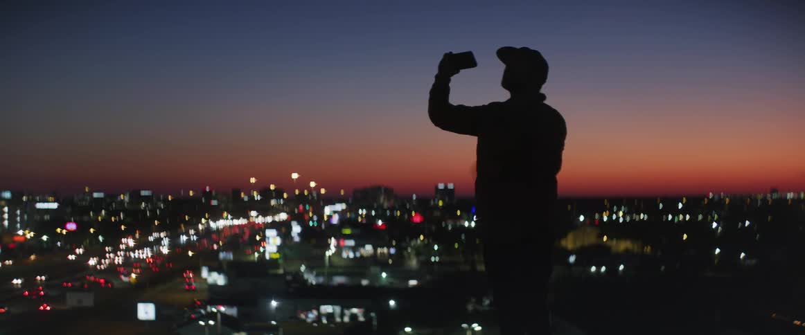 The silhouette of a man can be seen using his phone to take a photo of an illuminated city area