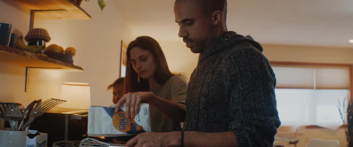 A man stirs food in a mixing bowl and a woman holding a toddler pours some flour in for him