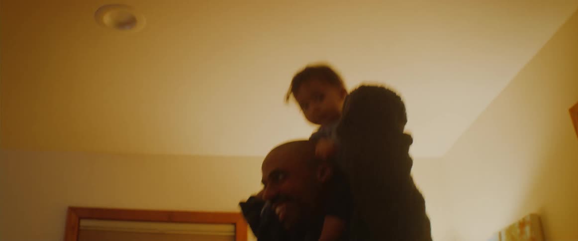 Man carries a baby on his shoulders and dances around the room while a woman watches from the sofa