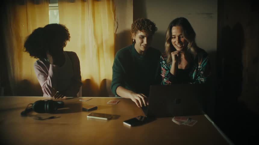Cellphones and headphones sit on a table as three women sit around it and look at a laptop and laugh