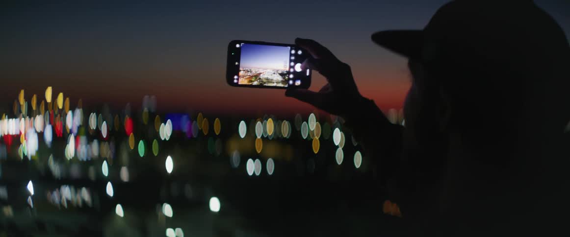 Silhouette of man taking pictures of city lights at night on cell phone