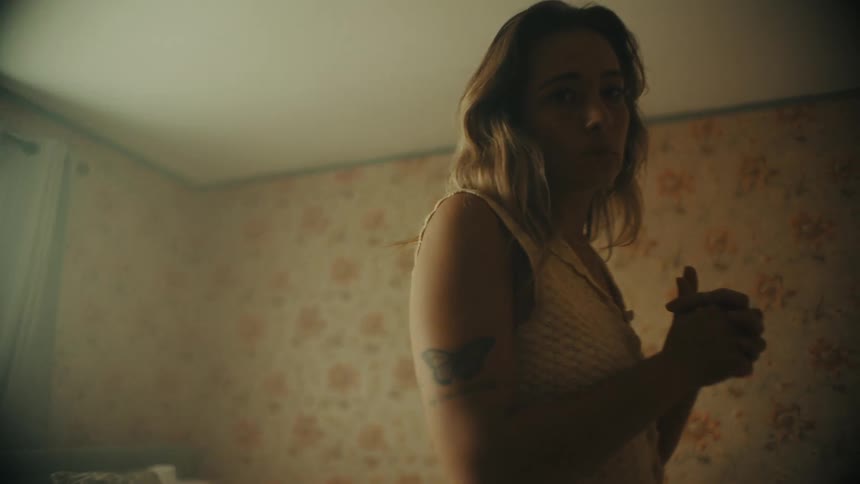 Blond woman with a butterfly tattoo walks across the floral wallpapered bedroom while clasping her hands