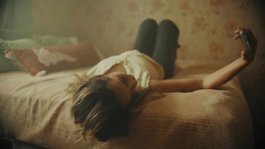 White woman lying in bed at her bedroom holding a smartphone posing for a selfie