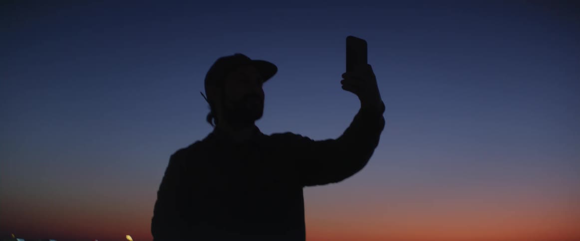 Man wearing a baseball cap using a smartphone to take selfies with the view of a big city in the background during sunset