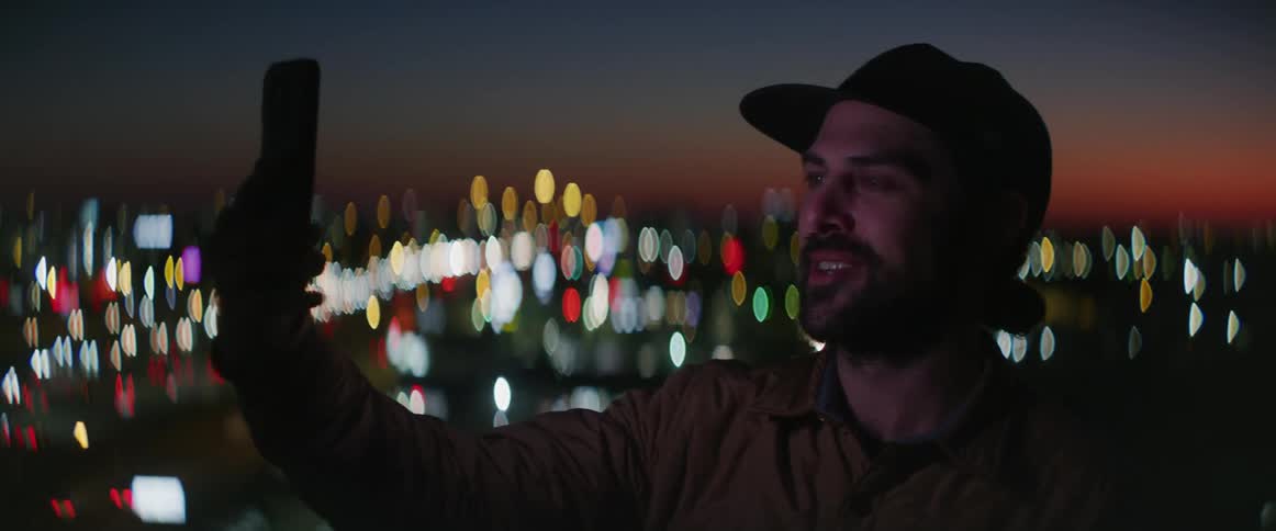 A man holds his phone up for a video call while standing outside overlooking downtown