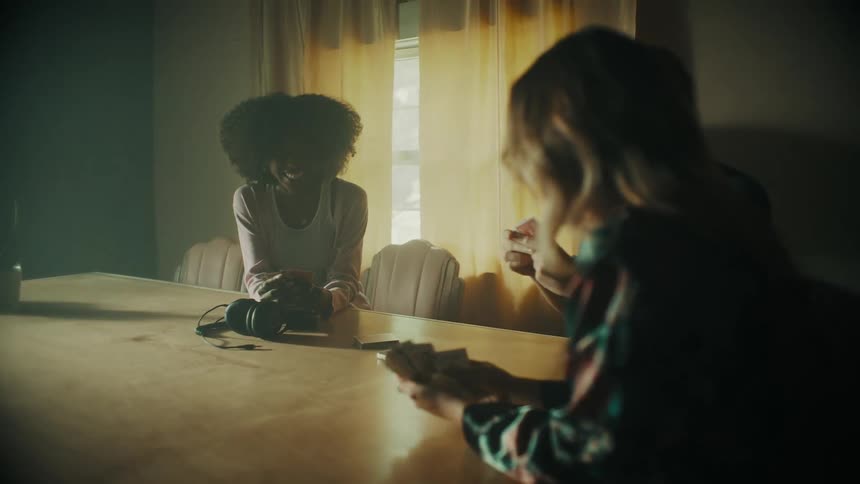 Three women sit down at a table and play cards as cellphones and headphones sit on the table