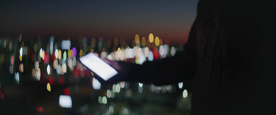Man with a beard raises his cell phone to look at the screen in the dark as traffic travels behind him