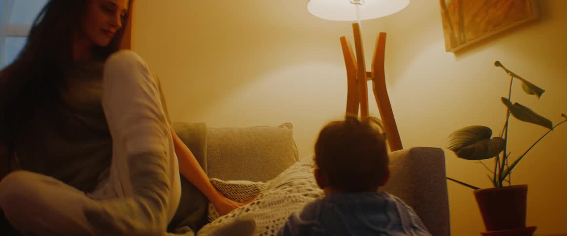 A woman encourages her son to climb onto the couch after he crawls over to her