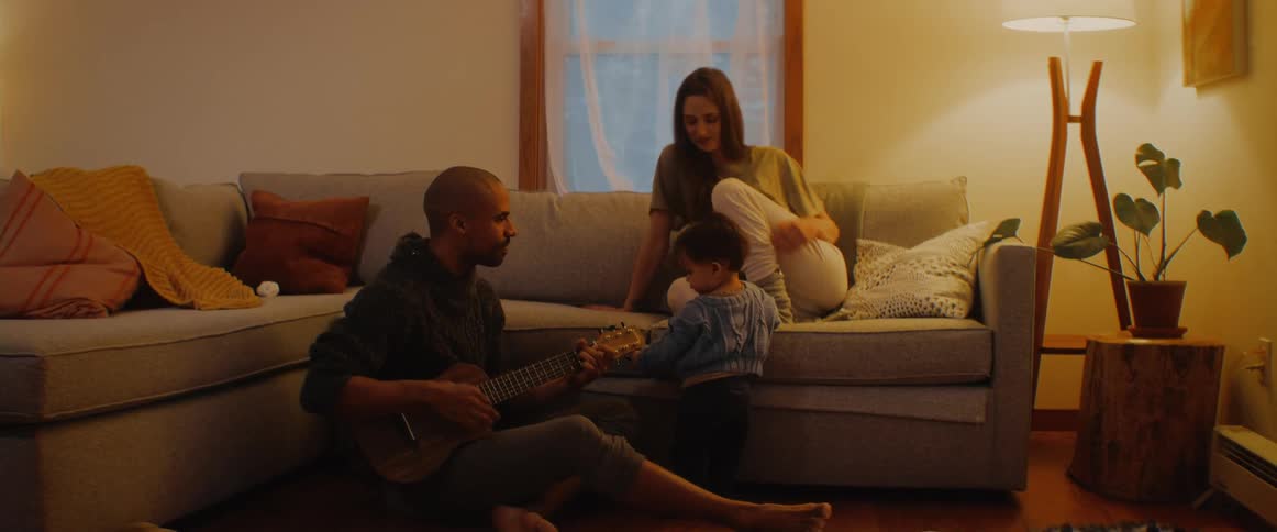 A man sits on the floor at home and plays the guitar and sings for his wife and son