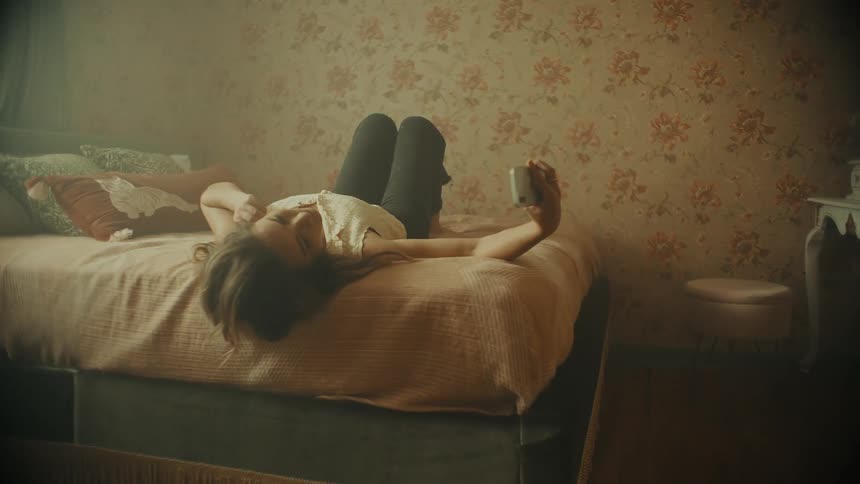 A teenage girl lies on her bed and holds her cell phone as she adjusts her clothing and hair before taking a selfie