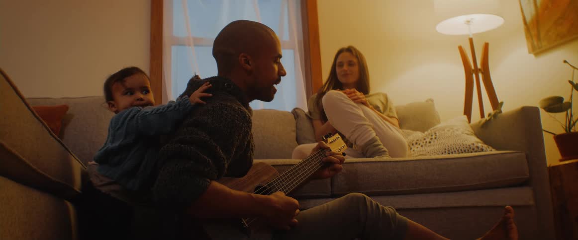 Mom looks on smiling from the couch while baby crawls all over daddy playing the guitar