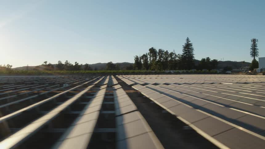 The sun is shining brightly on solar panels on a solar farm with trees and a building in the distance