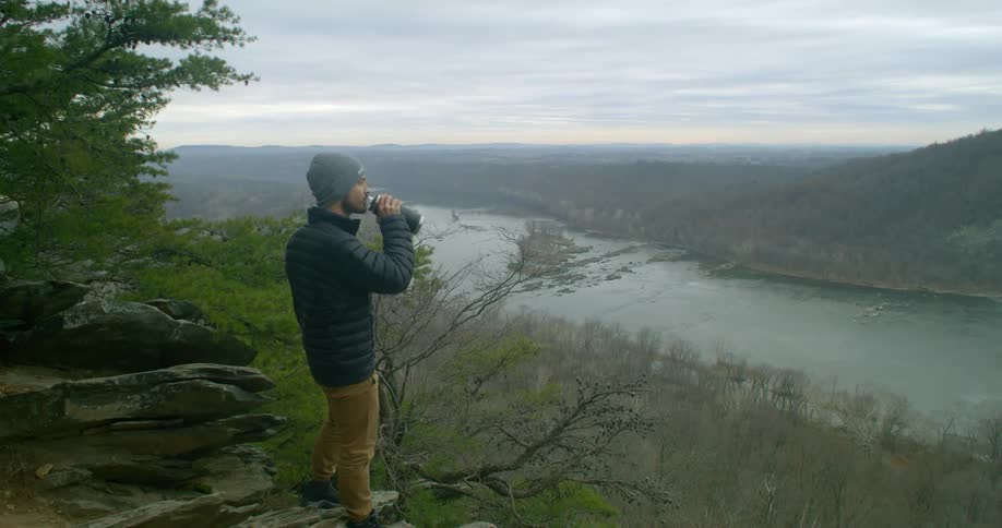 Young white man wearing a coat and winter cap drinking water from his water bottle standing in a cliff with a view of a river and a forest