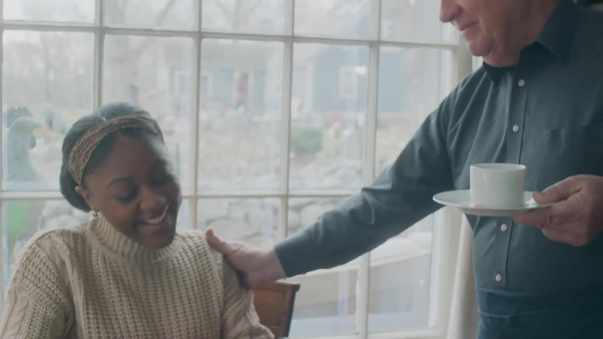 A young woman sits at a table in a home and an elderly man hands her a cup of coffee and pats her back