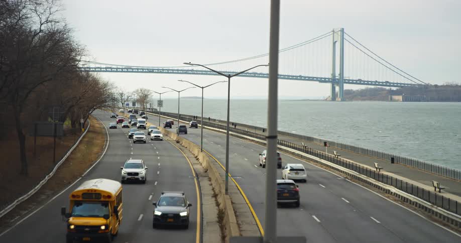 Vehicles drive on divided highway next to sea with suspension bridge in the background
