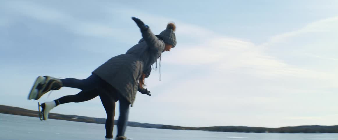Two young women hold hands and lift their legs up as they skate on a frozen lake