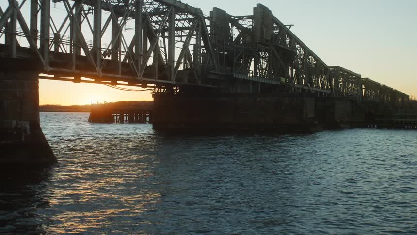 Sunlight reflects on the water as it flows under a bridge with a bright sun on the horizon