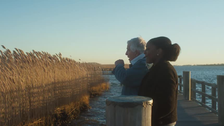 Young woman and elderly man stand on a pier at a lake and look at the reeds in the water