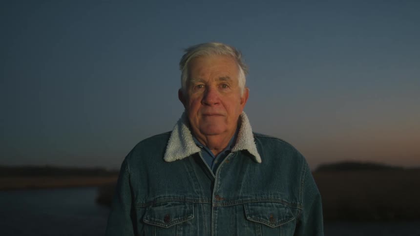 Elderly man with denim jacket faces the camera with eye contact at dusk
