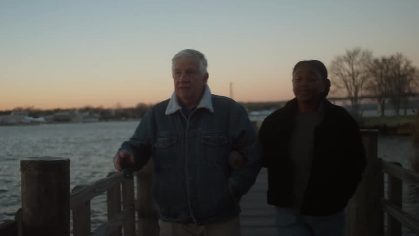 A young woman with her arm linked through an elderly mans arm walks along the pier as he talks with her