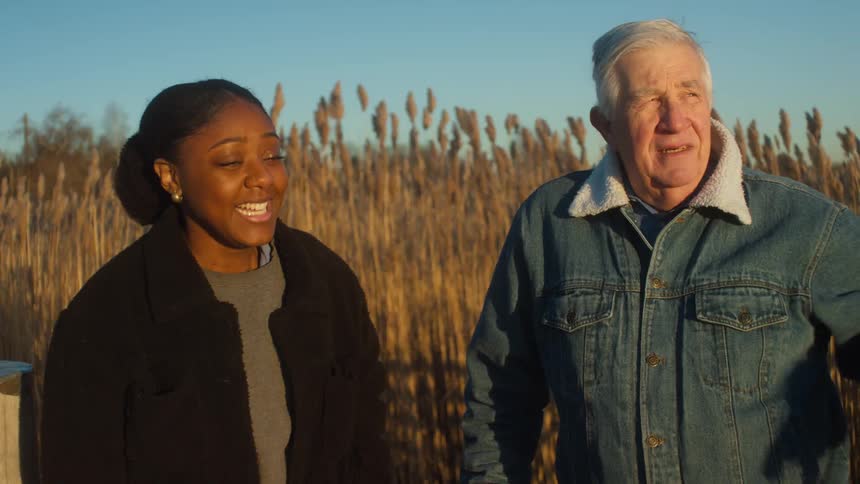 An elderly man and a young woman are standing outdoors in a rural area having a conversation on windy day at sunset
