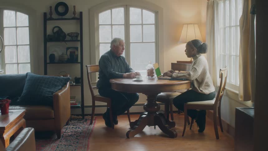 Old man and black woman sitting in the table next to the windows having a conversation while the woman is using a laptop