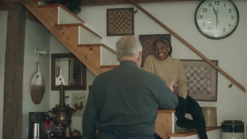 A woman sits on some stairs in a home and an elderly man walks over to her and takes her hand and helps her stand up and walk down