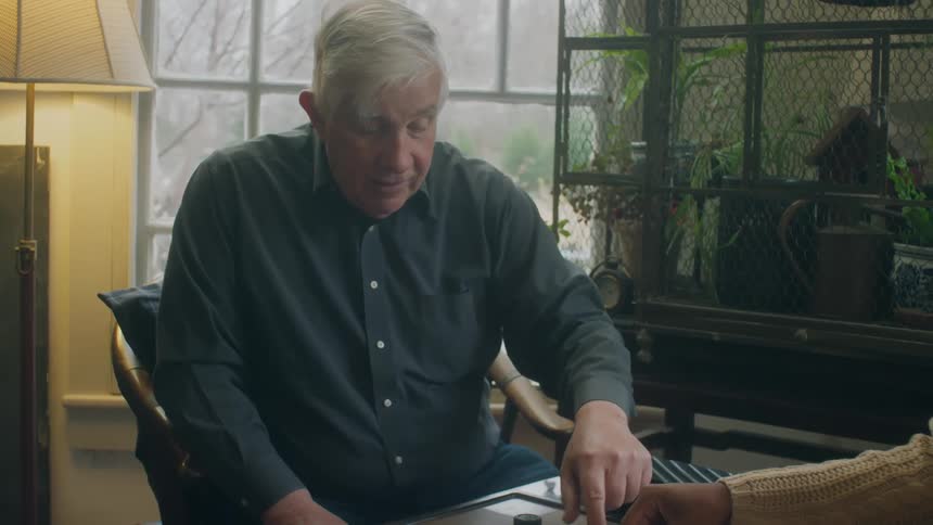 Elderly man sits playing checkers and talking near window showing winter scene