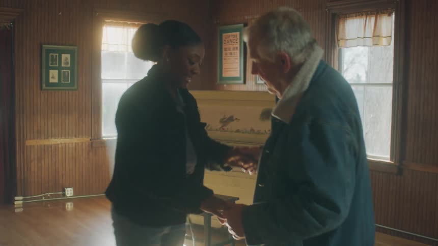 Elderly man wearing denim jacket and brown pants dances with woman with bun hair black coat and jacket next to piano in big room at home